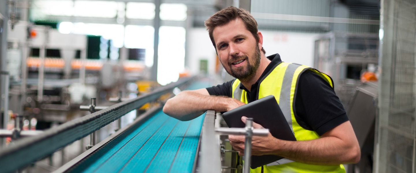 Lächelnder Mann in Warnweste und mit einem Tablet in der Hand steht in einer Produktionshalle.