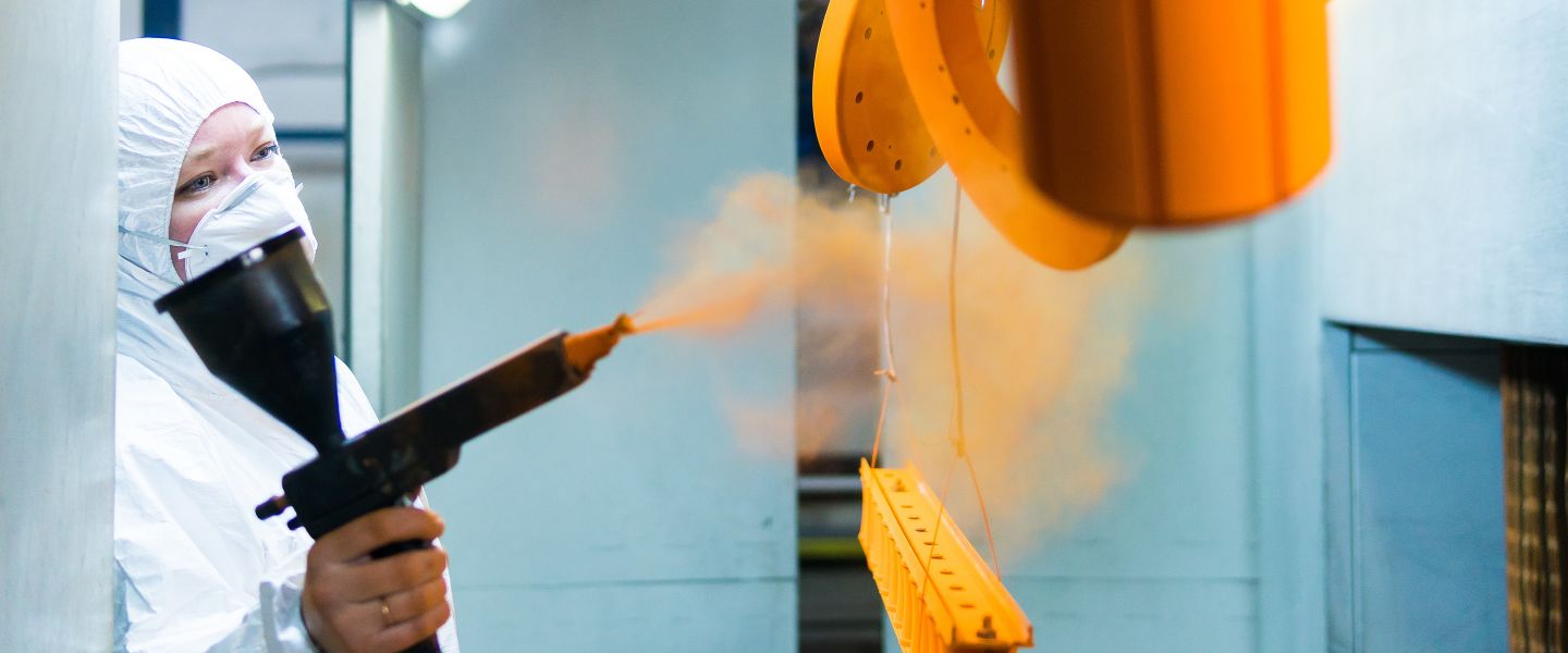 Person in Schutzkleidung bringt orangefarbenen Pulverlack mit Pulverpistole in der Pulverkabine auf.