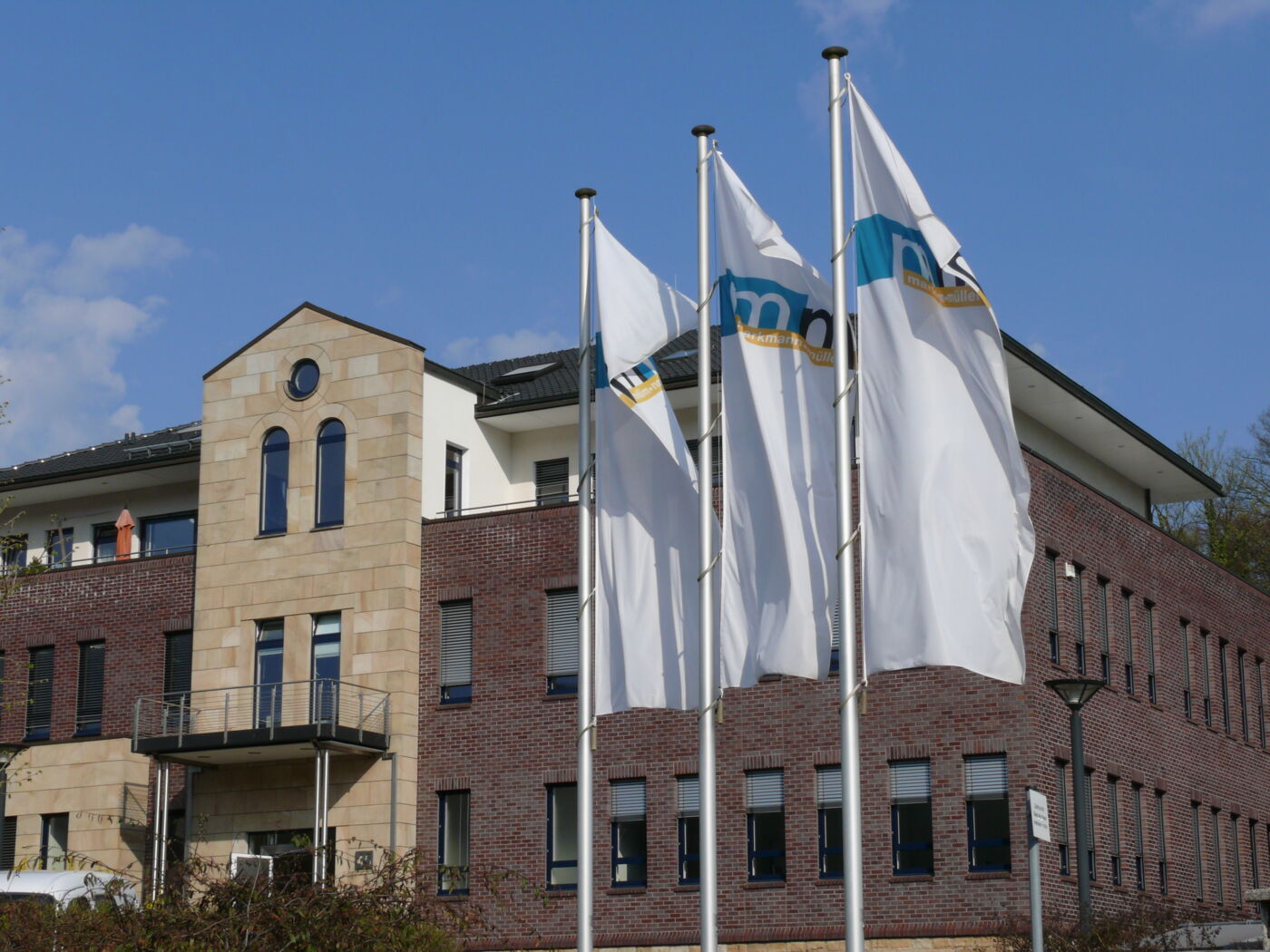 Blick von rechts auf den modernen Unternehmenssitz des Beratungs- und Software-Hauses markmann + müller in Herdecke. Im Vordergrund wehen drei weiße Fahnen mit dem Logo der Firma.